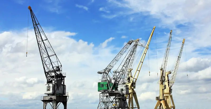 Several industrial cranes under a bright sky with scattered clouds